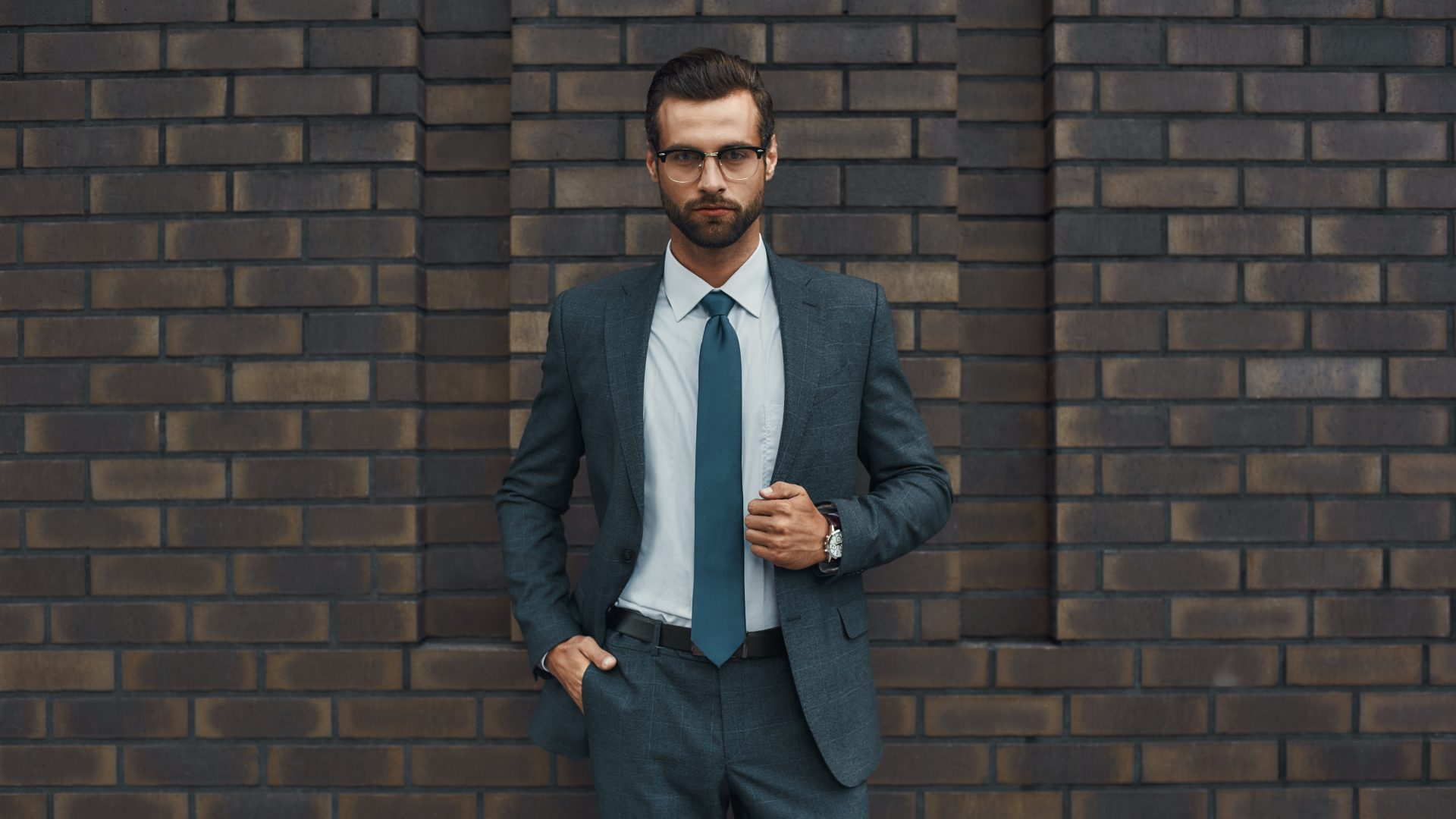 Great style. Confident businessman in full suit keeping hand in pocket and looking at camera while standing against brick wall